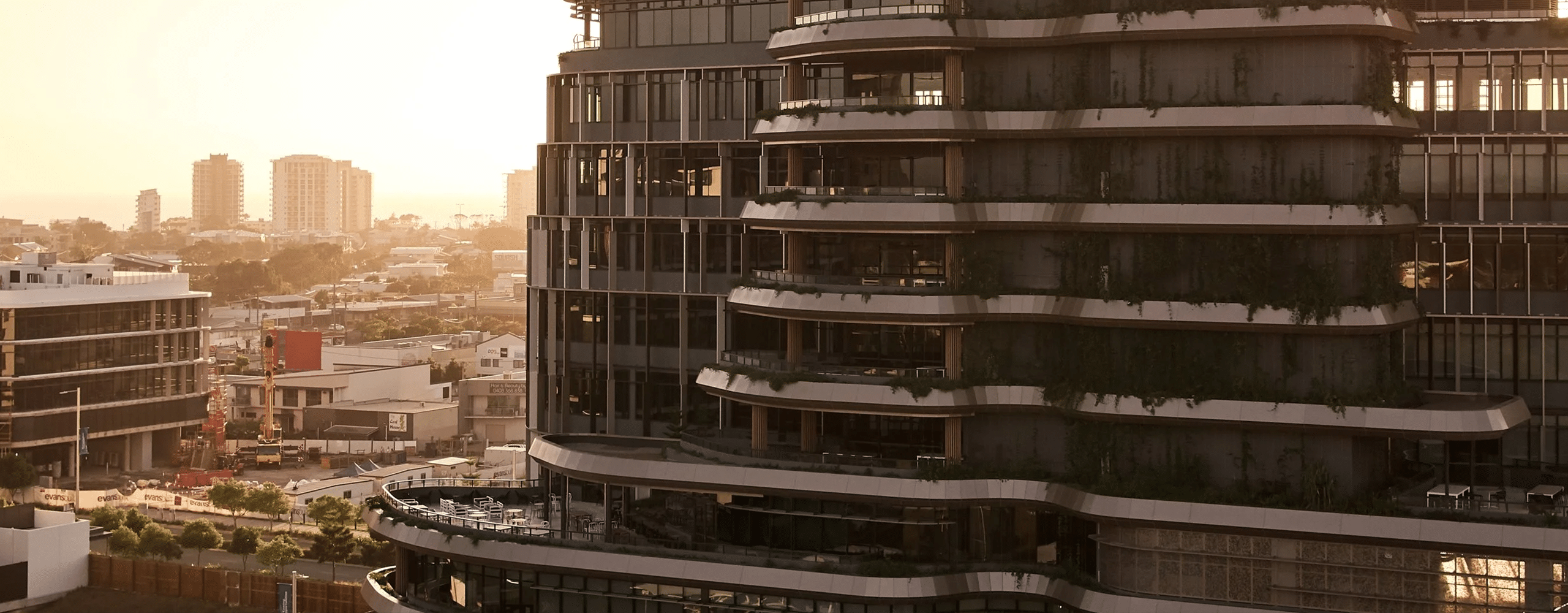 A contemporary curved tower with planted balconies in the Maroochydore CBD, with the surrounding suburb visible at sunset.