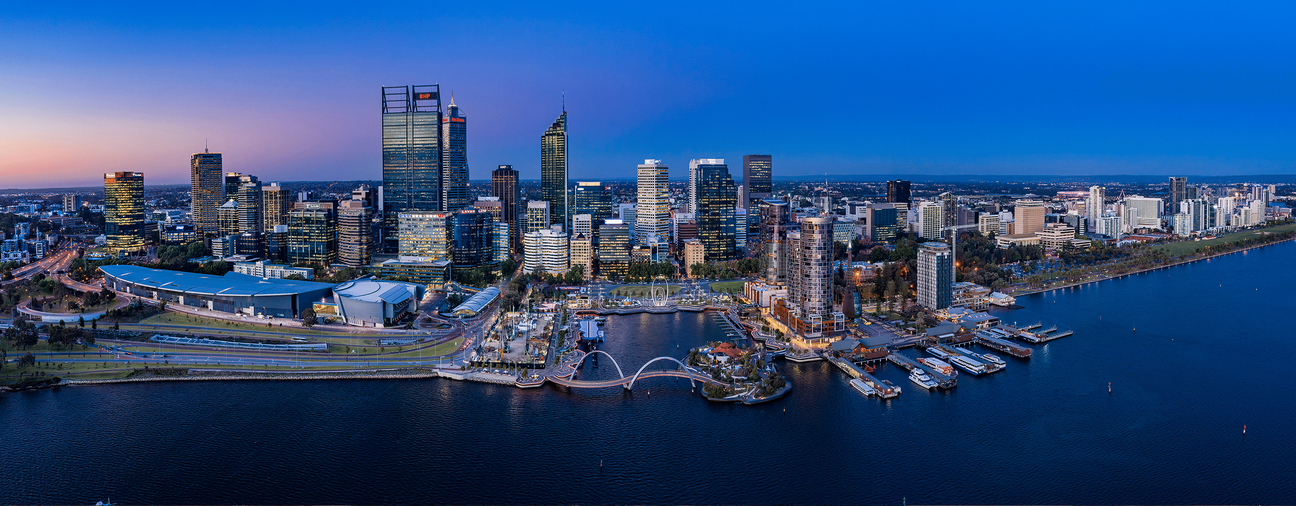 Panoramic aerial view of Perth CBD and Elizabeth Quay at dusk, with the Swan River in the foreground and the city skyline lit against a deep blue sky.