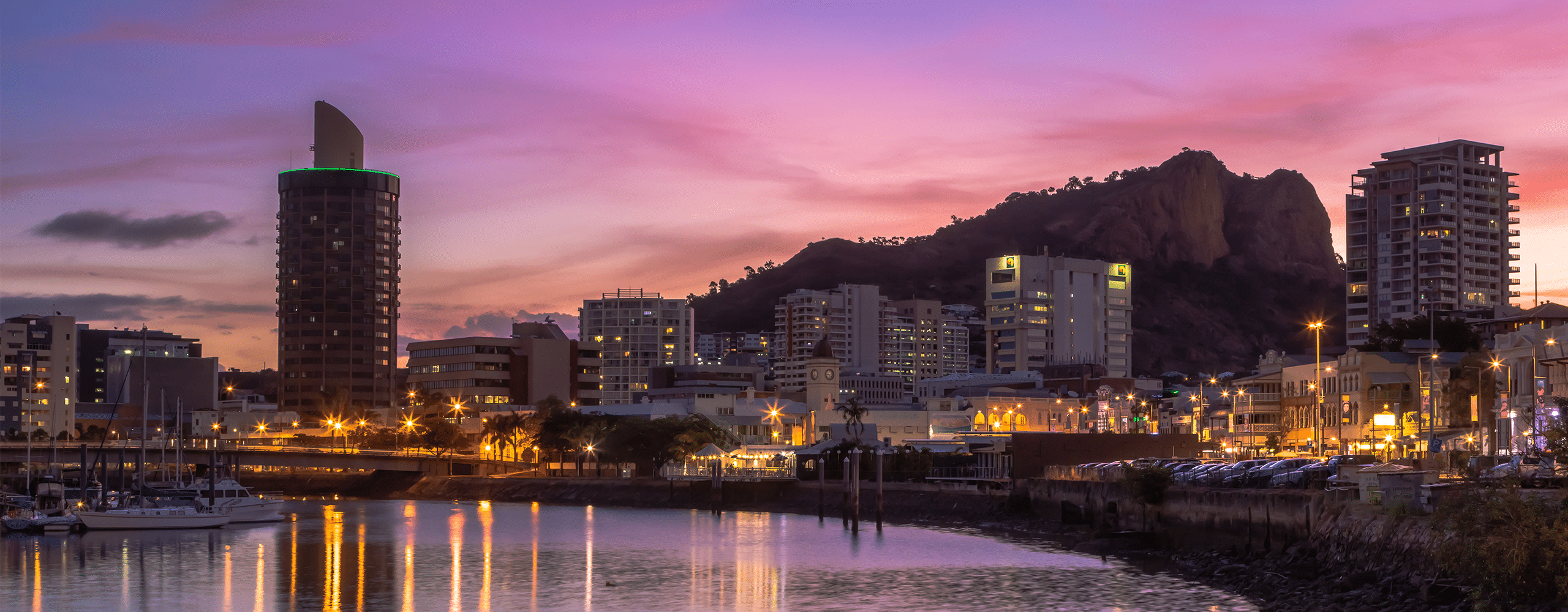 Townsville city skyline and waterfront at dusk, with Castle Hill in the background and warm street lights reflecting on the harbour.